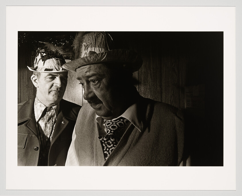Black and white photograph of two men wearing party hats marked "Happy New Year." The man in the foreground appears pensive and is looking down, while the man in the background is looking off to the side with a serious expression. Both are dressed in suits and the setting suggests an indoor environment with a wooden wall behind them.