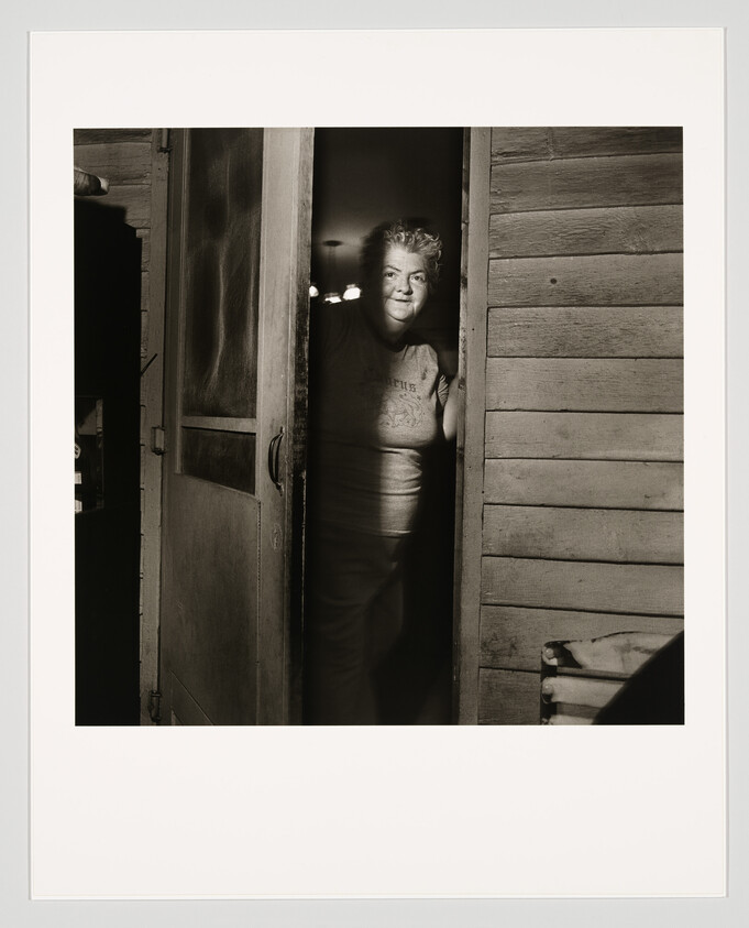 A black and white photograph of a person standing in a doorway, partially illuminated by light coming from inside the room. The individual is smiling and looking directly at the camera. The wooden texture of the door and the adjacent wall is visible, adding a rustic feel to the image.