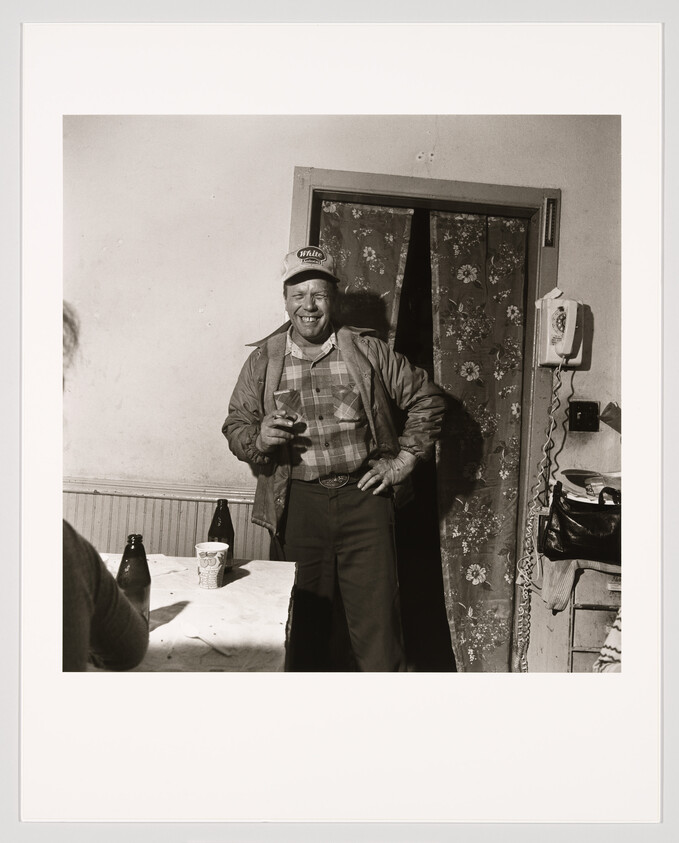A black and white photograph of a smiling man standing by a door, wearing a cap, plaid shirt, and jacket, holding a glass in one hand with beer bottles on a table nearby.