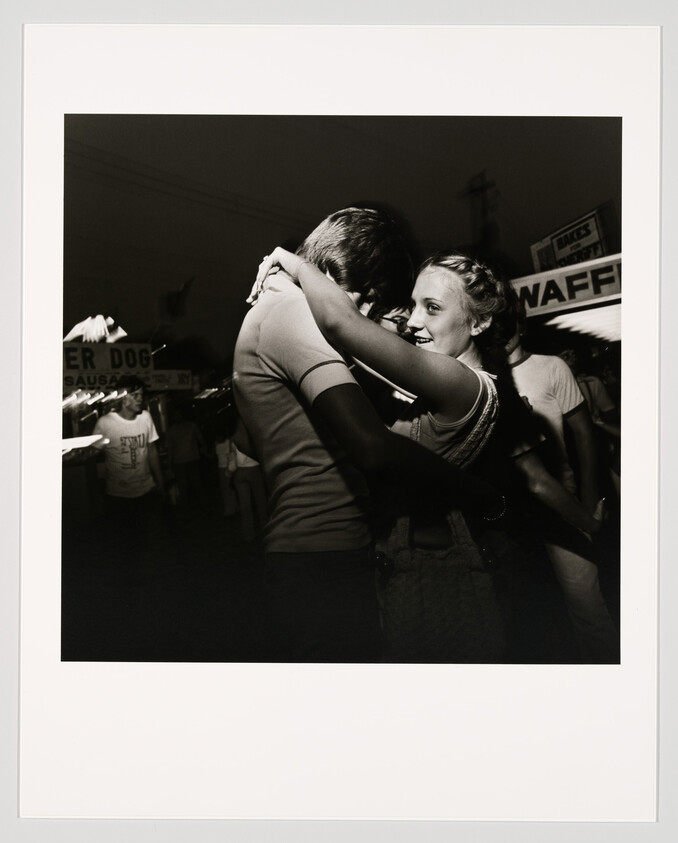 A black and white photograph capturing a tender moment where a couple is embracing and dancing, with the woman smiling and looking at the man affectionately. They are surrounded by a bustling fairground atmosphere with signs for food stands like "WAFFLES" and "SUPER DOG" in the background.
