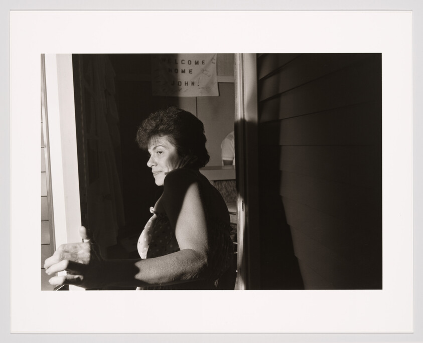 A black and white photograph of a woman sitting in a doorway, with sunlight casting half of her face in shadow. She has a contemplative expression and is looking to the side. Behind her, a sign reads "WELCOME HOME JOHN." The contrast of light and shadow creates a dramatic effect.