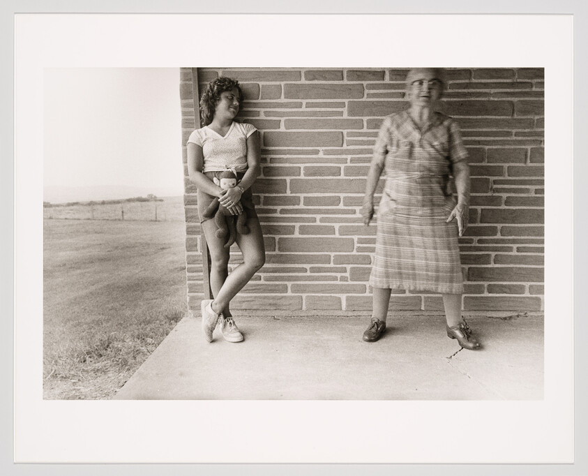 A black and white photograph showing two people against a brick wall. On the left, a young woman leans against the wall, looking to the side, holding a small stuffed animal, and wearing a short-sleeved top, shorts, and sneakers. On the right, an older woman appears blurred due to motion, standing with her hands on her hips, wearing a plaid dress and loafers. The background features an open field and a clear sky.