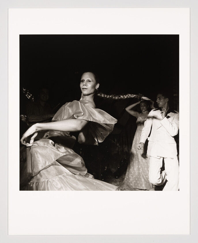 A black and white photograph capturing a moment at a dance, featuring a woman in a flowing dress with her arm extended gracefully and a man in a white suit dancing behind her. The woman's hair is styled in a long braid that swings behind her, and the lighting highlights the dynamic movement of the dancers.