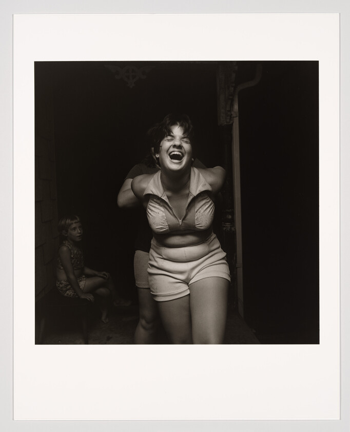 A young woman laughs joyfully as someone playfully holds her arms from behind on a porch.