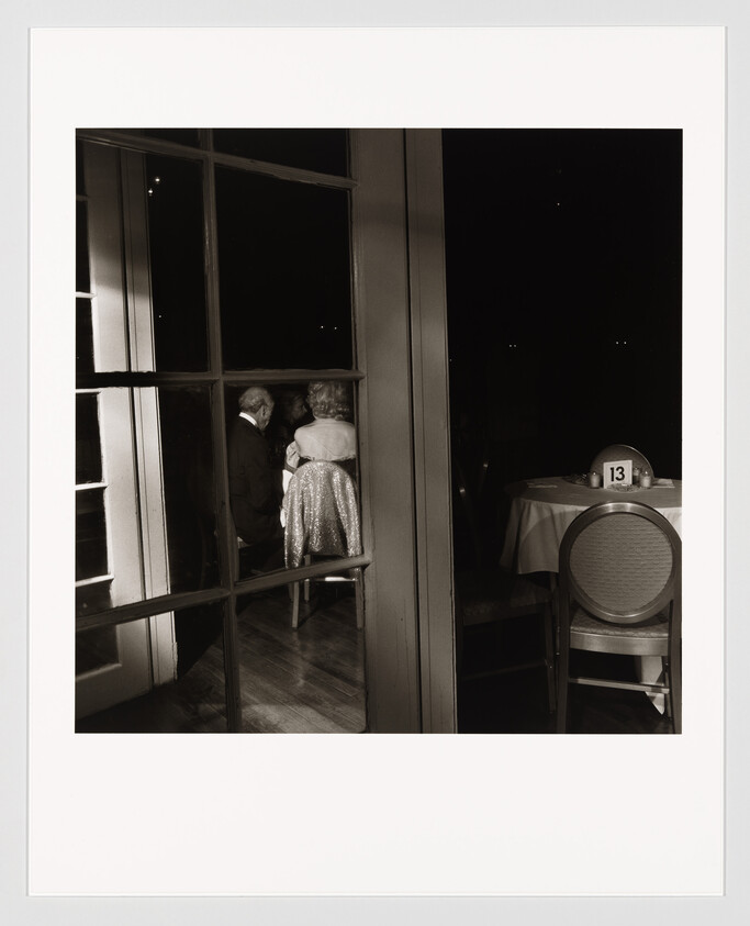 An elderly couple sits close together by a window in a dim dining room, table marked 13.
