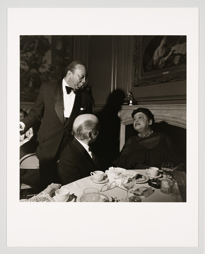 A man in a tuxedo leans forward speaking to a seated woman and man at a dinner table.