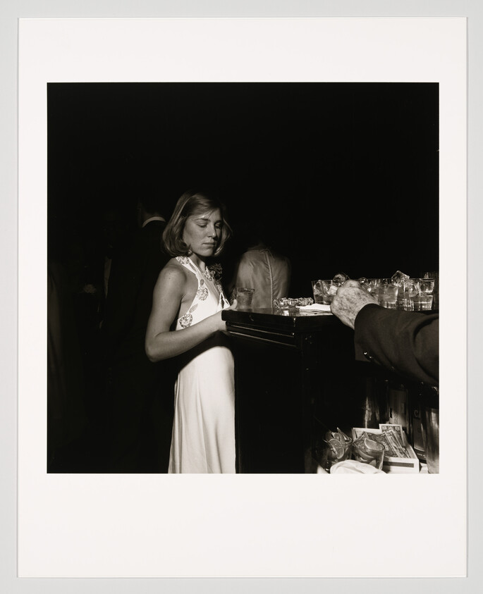 A woman in a white dress stands at a dim bar looking down as drinks are served.