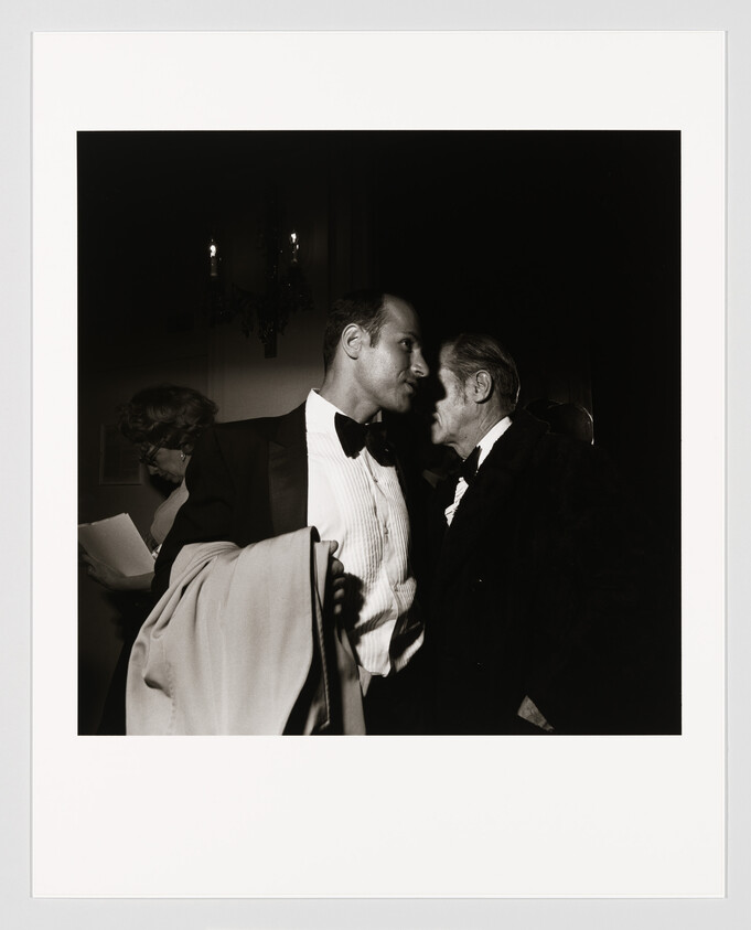 Two men in tuxedos lean together whispering closely at a formal event while one holds a coat.