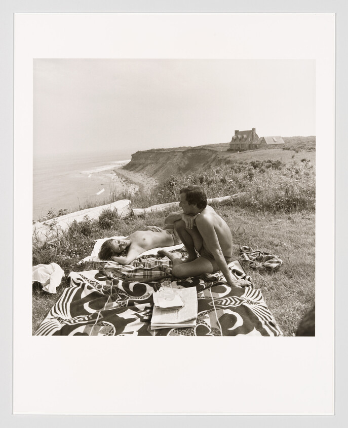 Two people relax on a blanket by a coastal cliff, with a house visible in the distance.