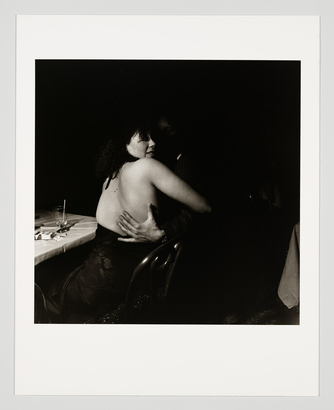 Woman in a backless dress sitting at a table embraces a person in a dimly lit room.