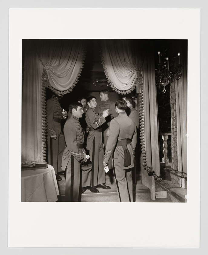 A group of young men in dress military uniforms converse and gather beneath ornate draped curtains.