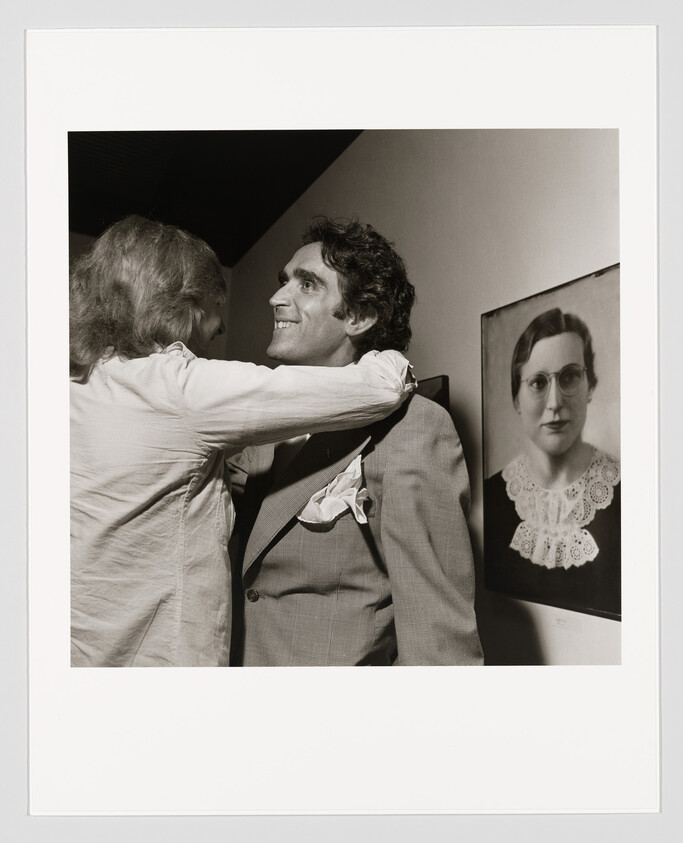 A man embraces a smiling man in a suit beside a framed portrait of a woman.
