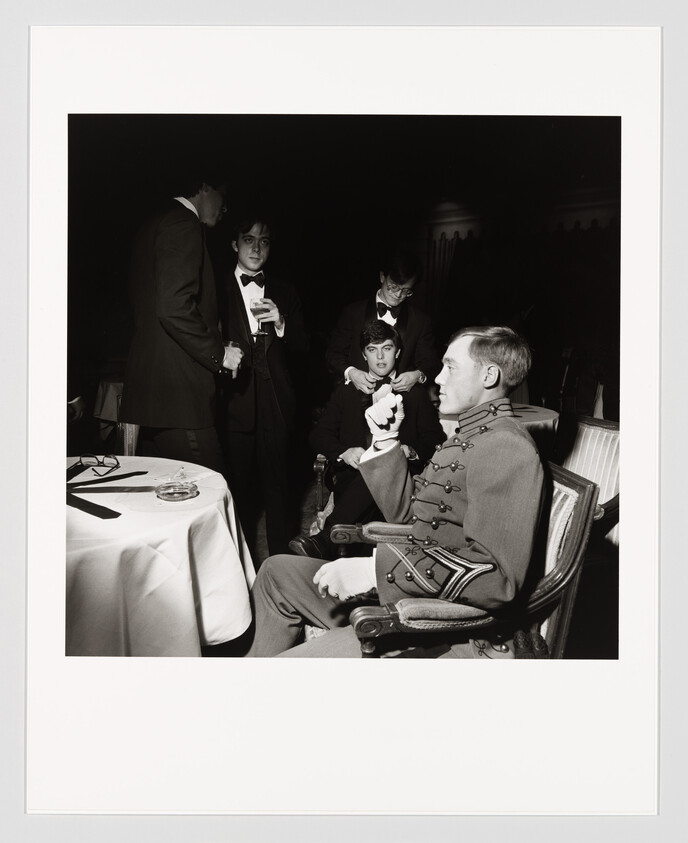 A young man in a military uniform sits while three men in tuxedos stand and adjust each other's bow ties.