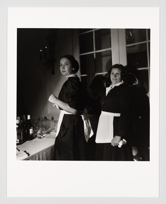Two women in maid uniforms stand beside a table of bottles and glasses, looking toward the camera.
