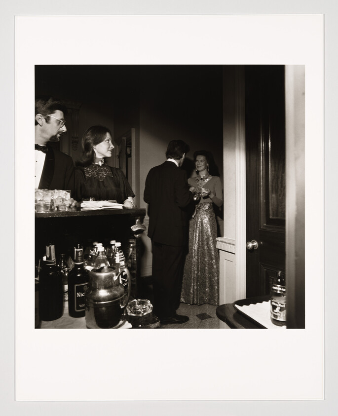A black and white photograph capturing a moment at a social gathering, with a man in a suit and a woman in a dark dress standing by a bar with various bottles, looking towards another couple engaged in conversation. The second woman is wearing a sparkling dress and holding a drink, with an open door in the background suggesting an adjoining room. The scene has a vintage feel, reminiscent of mid-20th century parties.