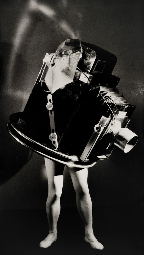 Person stands wearing a large mechanical camera-like contraption covering their head and torso.