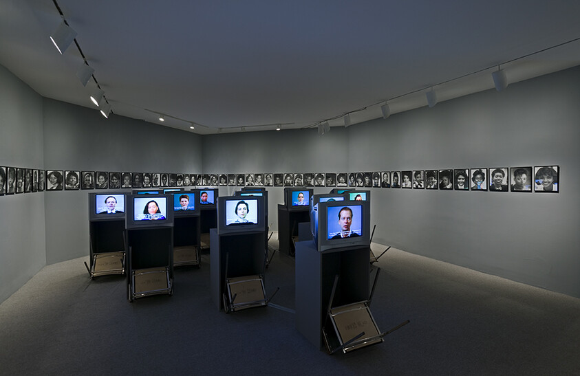 Multiple vintage televisions on stands display talking faces while a wall of black-and-white portraits surrounds them.