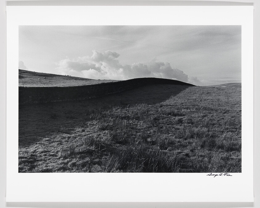 Black and white photo of rolling hills with textured grass and a dramatic sky, signed by the artist in the lower right corner.