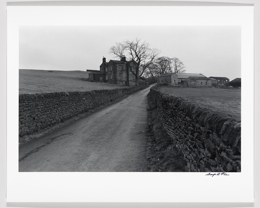 A black and white photograph depicting a rural scene with a country road flanked by a stone wall leading towards a traditional stone-built house and outbuildings, with leafless trees and a barren landscape in the background. There is a signature on the bottom right corner of the image.