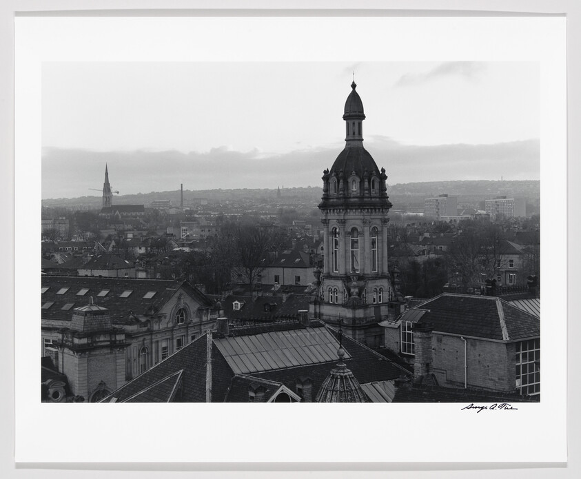 A black and white photograph capturing a cityscape with a prominent, ornate tower in the foreground, surrounded by various rooftops, and a hazy skyline in the distance. The image is signed by the photographer in the lower right corner.