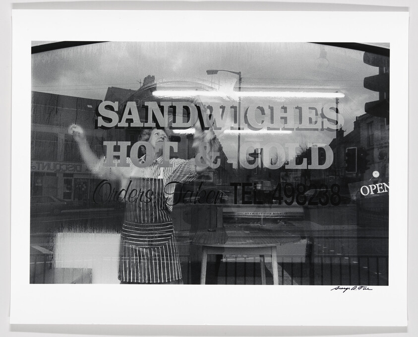Black and white photo of a person behind a window with "SANDWICHES HOT & COLD" text, reflecting cityscape.