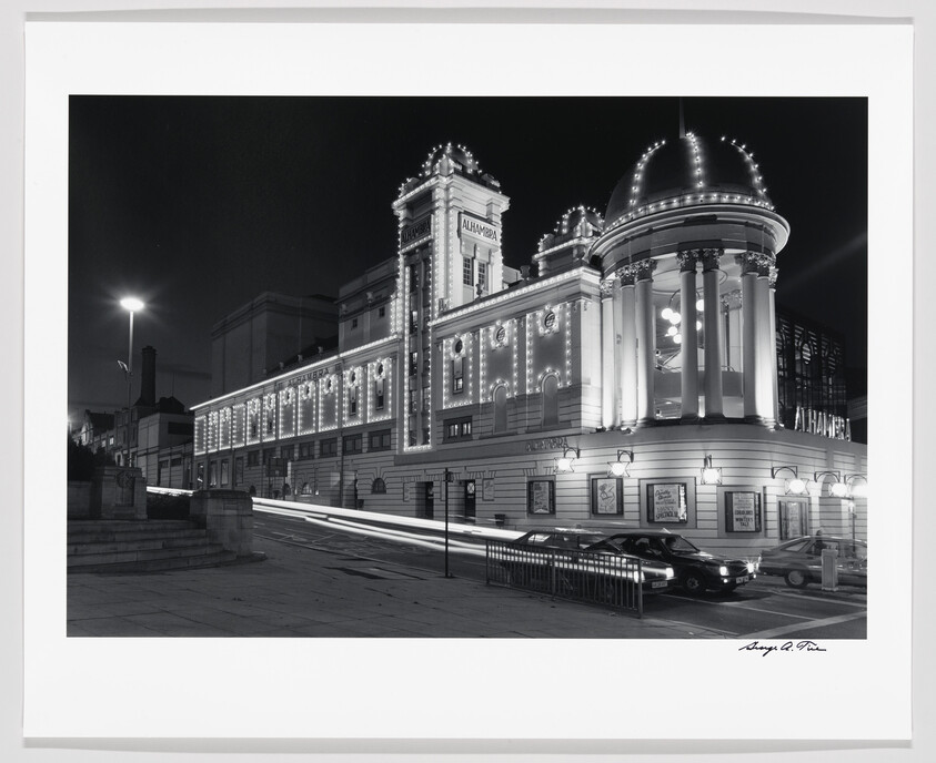 Black and white photo of the illuminated Alhambra Theatre at night with light trails from passing cars.