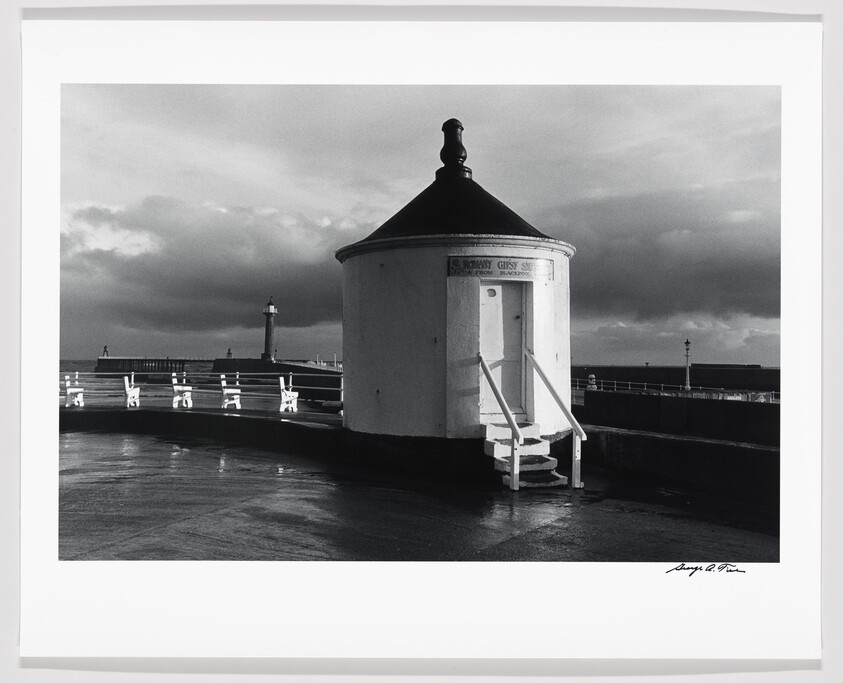 Black and white photo of a round building on a pier with a lighthouse in the background under a cloudy sky.