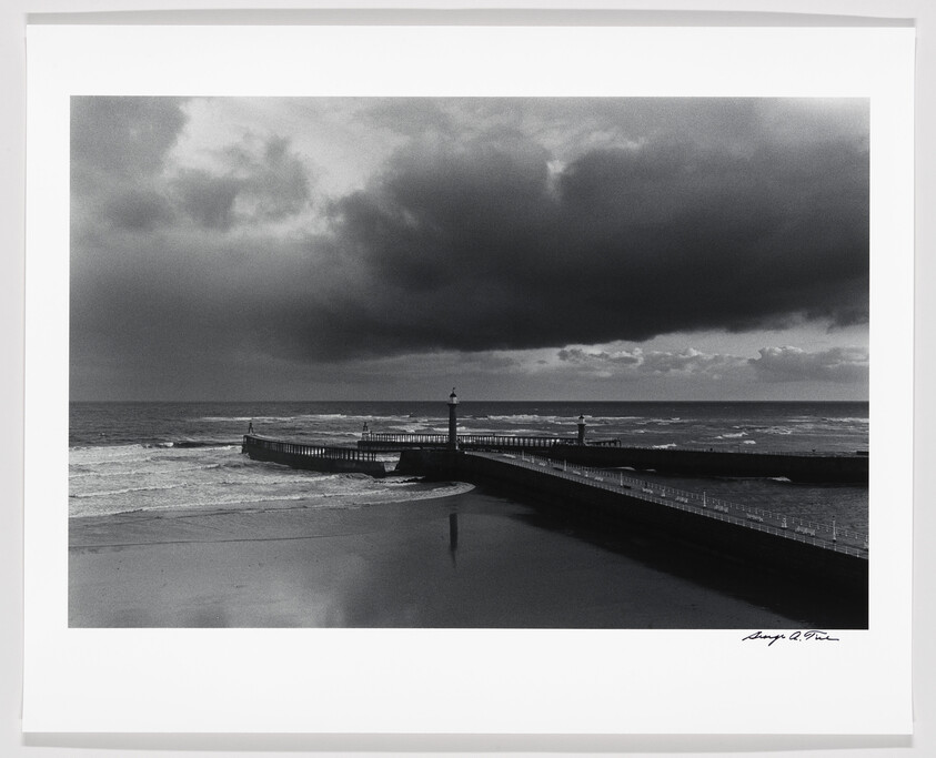 A pier with two lighthouses stretches into the rough sea under heavy storm clouds.