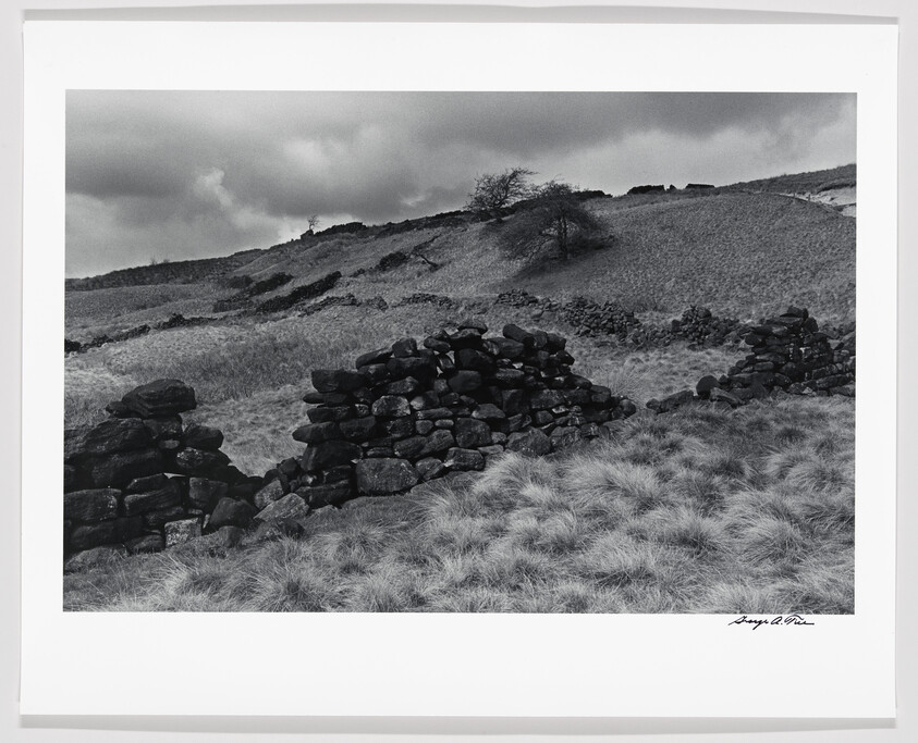 Black and white photo of a hilly landscape with a stone wall and grassy terrain under a cloudy sky, signed by Ansel Adams.