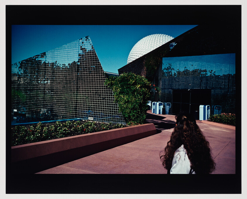 A person with long hair walks toward reflective glass buildings and a white geodesic dome.