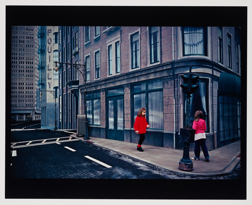 Two girls on a quiet city sidewalk near a traffic light, one in a red coat walking toward the other.