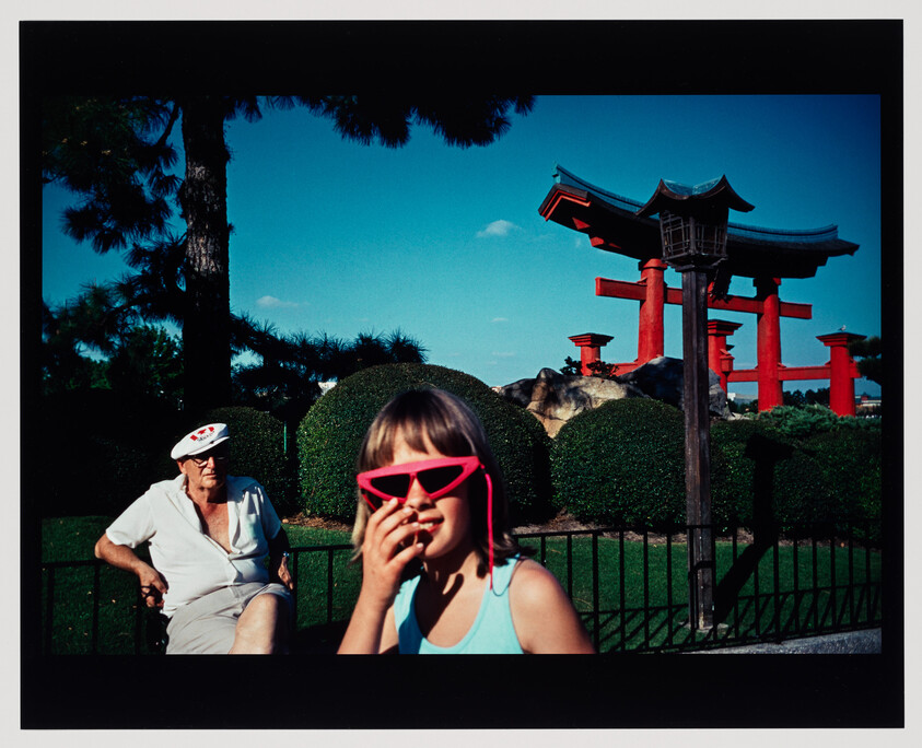 A girl wearing bright pink sunglasses covers her mouth while an older man sits near a red torii gate.