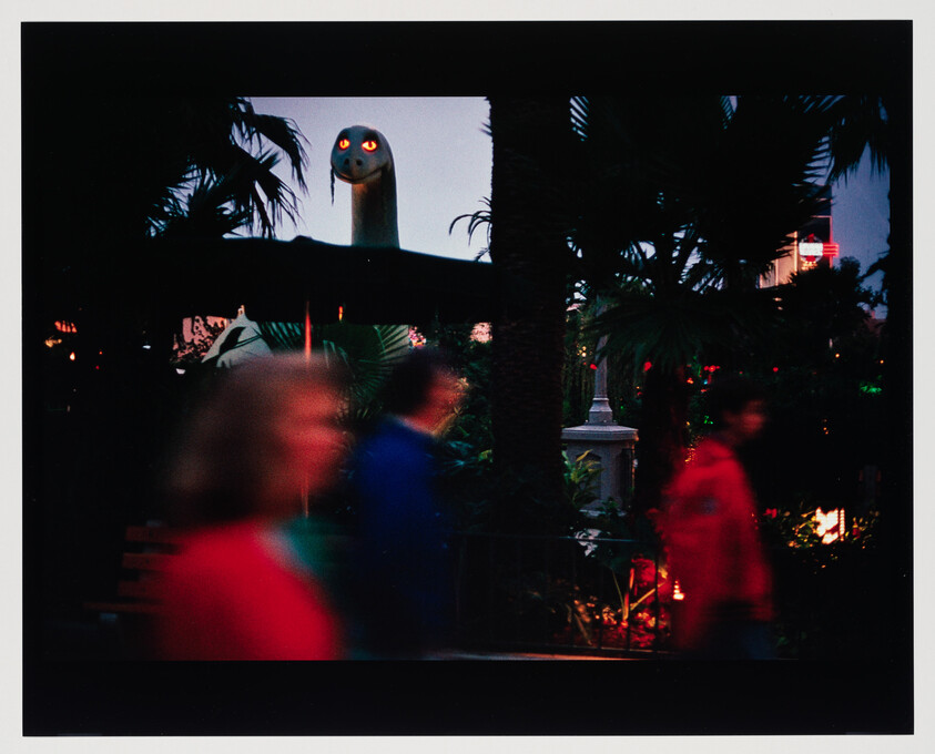 People walk past a dark amusement area while a large serpent head with glowing eyes watches above.