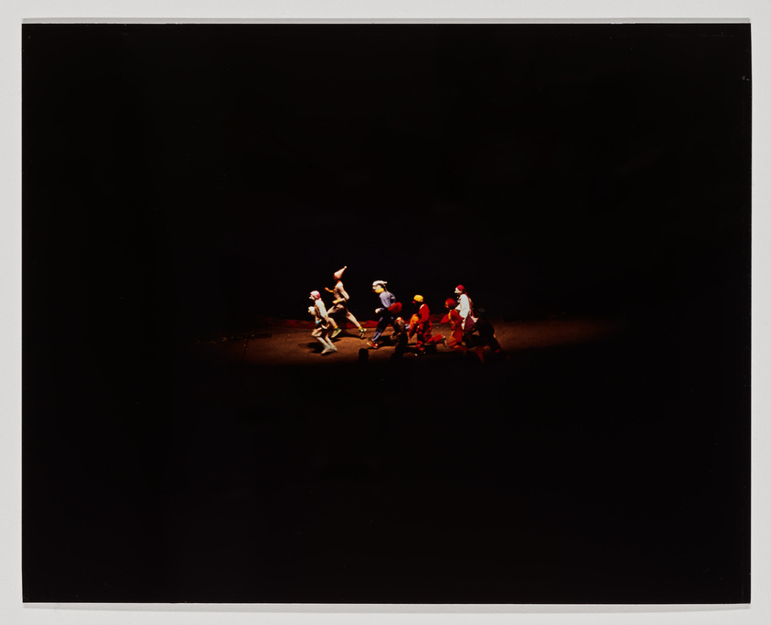 A group of performers on a dark stage illuminated by a spotlight, with some dressed in white and others in red, seemingly in motion as if part of a choreographed dance or theatrical performance.