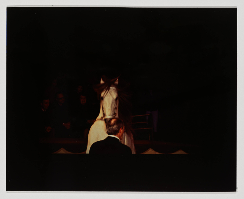 A photograph capturing a dramatic moment on stage with a person in a wide-brimmed hat and trench coat standing with their back to the camera, facing another person who is seated and facing away from the camera. The audience is visible in the background, watching the scene unfold in a dimly lit setting.