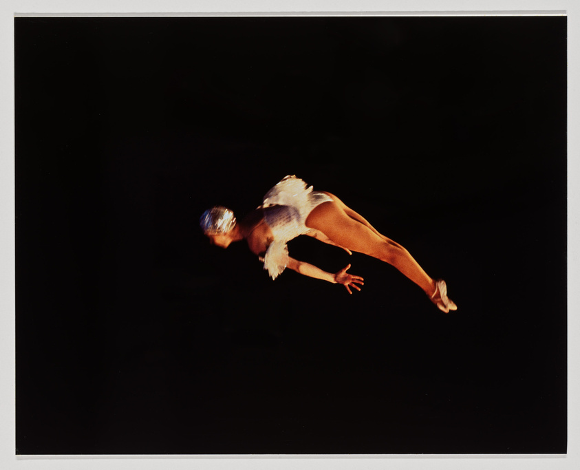 A ballet dancer captured mid-leap against a dark background, wearing a white costume with feathers and a sparkling headpiece.