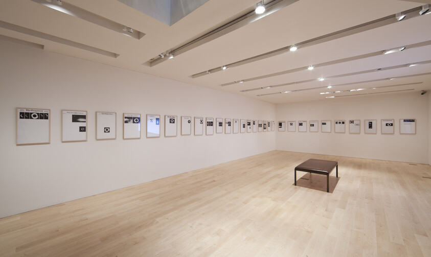 A spacious gallery with a long row of framed minimalist prints along the walls and a bench.