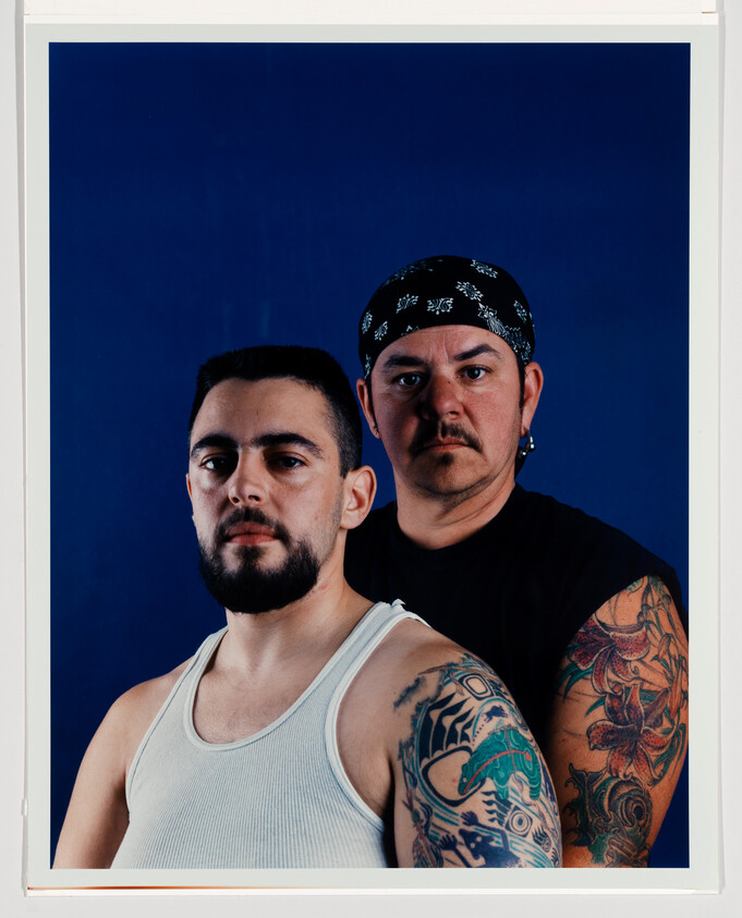 Two tattooed men stand together against a blue background, one wearing a bandana and tank top.