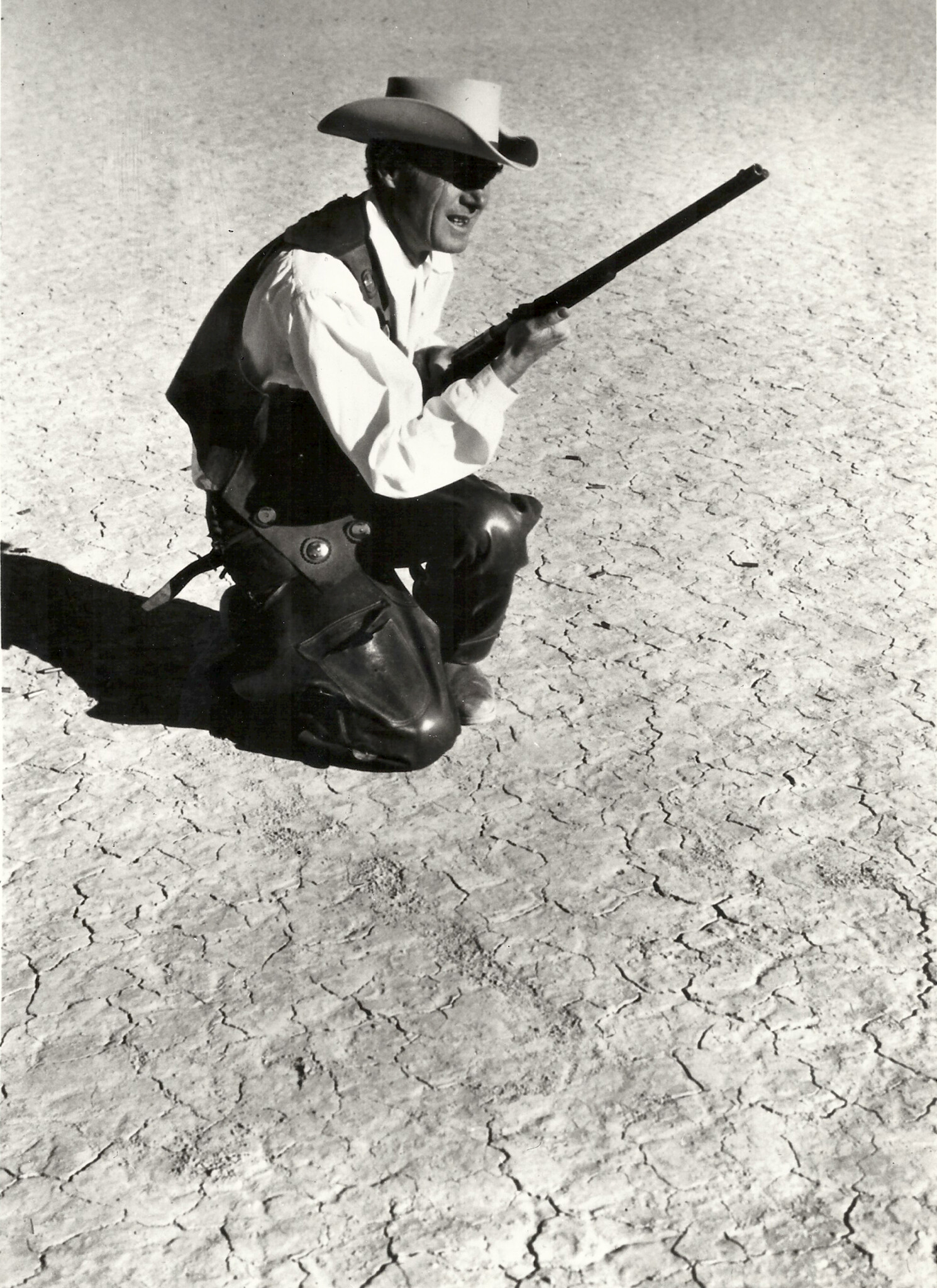 Black and white film still of a man in Western attire kneeling with a rifle on a cracked desert terrain