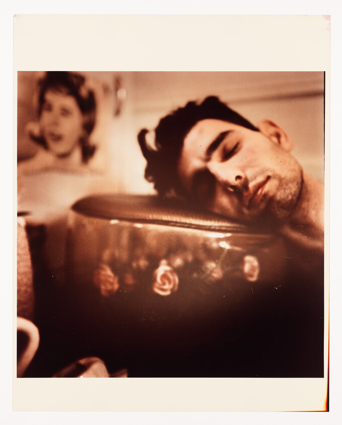 A young man sleeps with his head resting on a floral-patterned suitcase in a dim room.