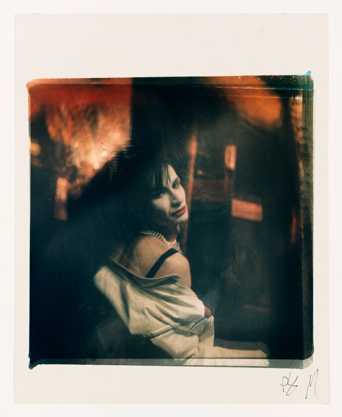 A woman with dark hair and a white jacket looks over her shoulder in a dimly lit, blurred setting.