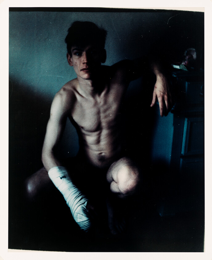 Shirtless young person crouches in dim light with a heavily bandaged forearm.
