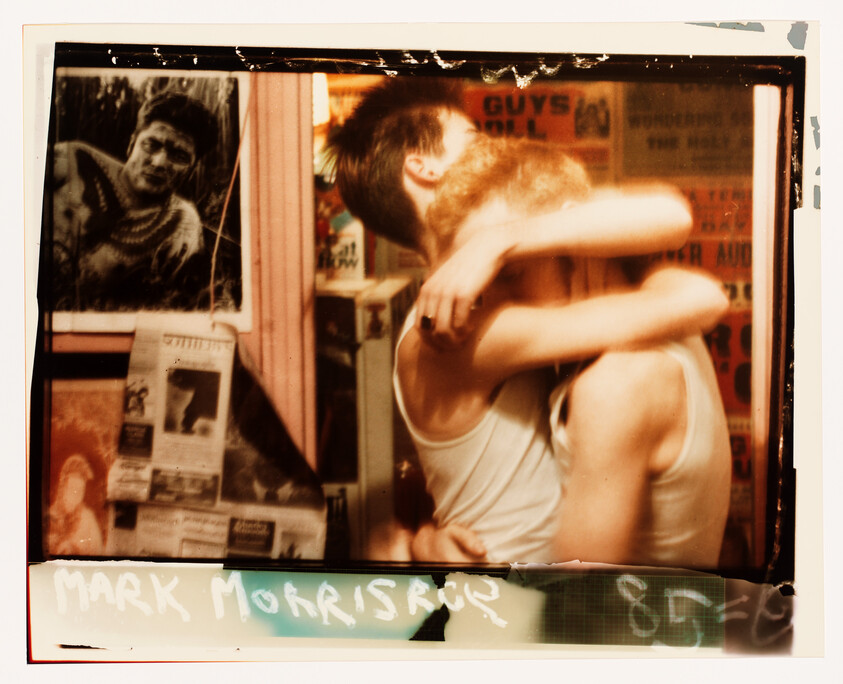 Two young men in white tank tops embrace and kiss in a poster-covered room.