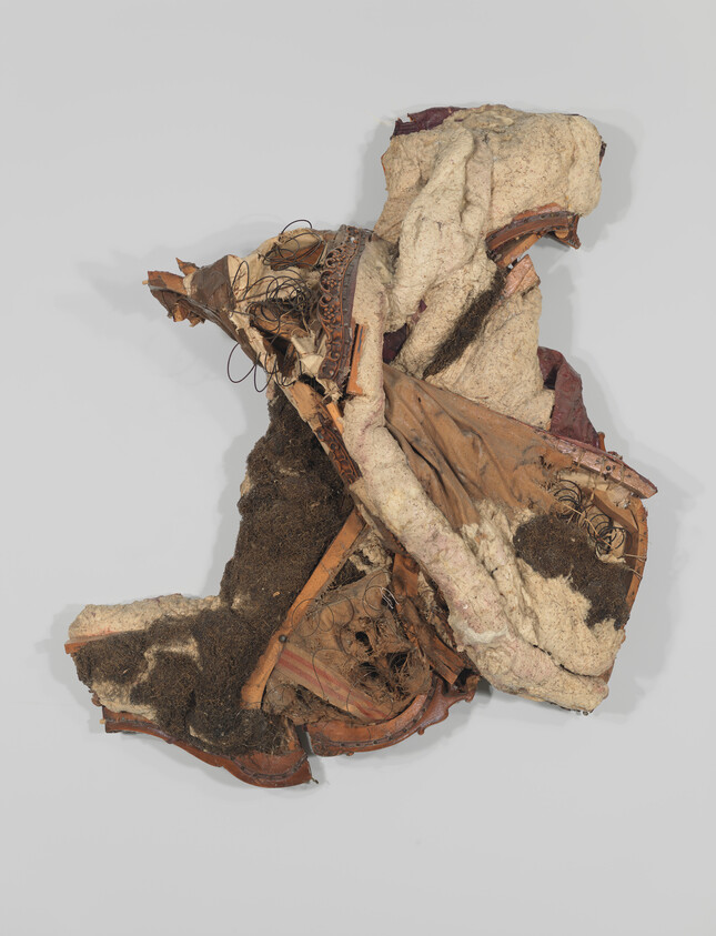 Torn upholstered chair seat with exposed springs, stuffing, and wooden frame arranged against a plain background.
