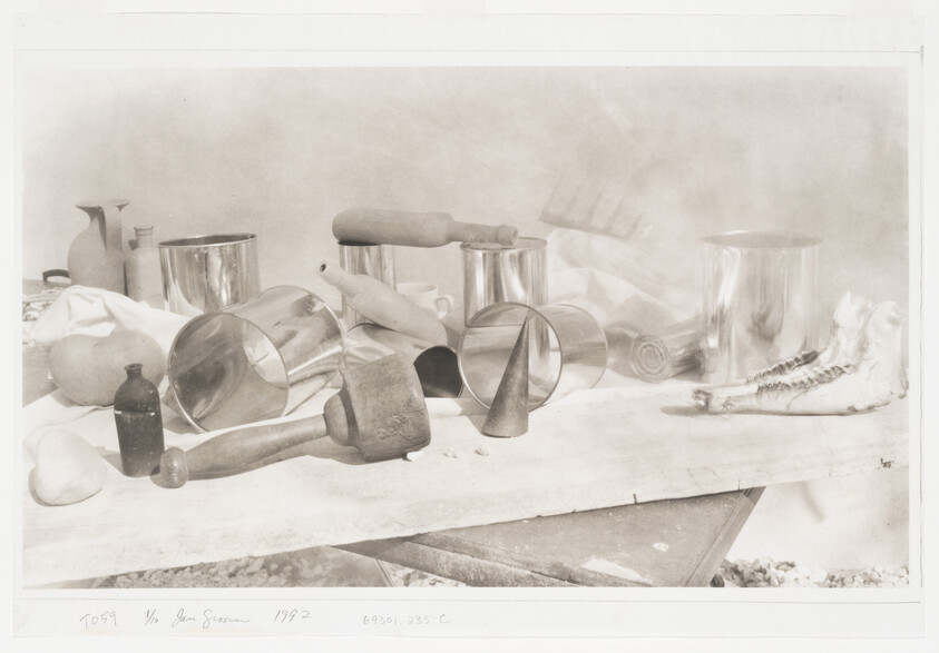 A still-life arrangement of kitchen tools, metal cups, a mallet, rolling pins, and a jawbone on a table.