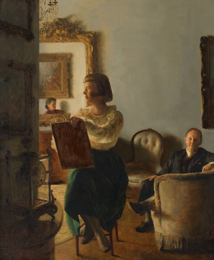 A woman sits holding a sketchbook while a man watches from a nearby armchair.