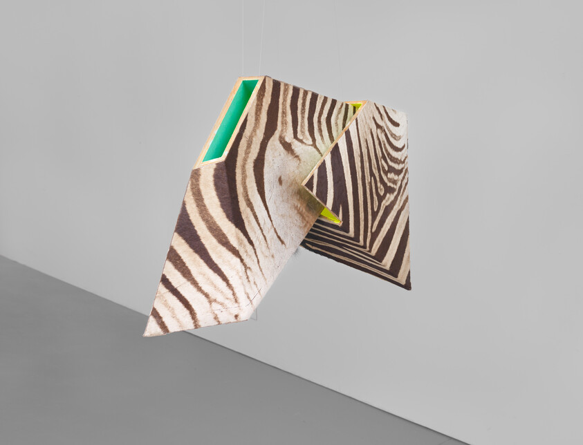 A suspended geometric sculpture with zebra stripes and neon green interior panels hangs in a gray room.