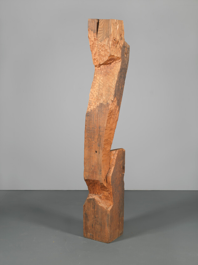 Tall rough-hewn wooden post with angular carved notches standing upright on a gray floor.