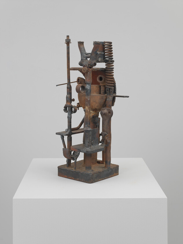 Abstract metal sculpture made from repurposed machine parts mounted on a white pedestal.