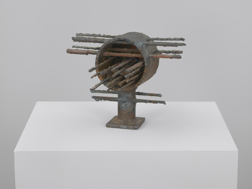 A rusted metal sculpture with a hollow cylinder and many rods protruding, on a pedestal.